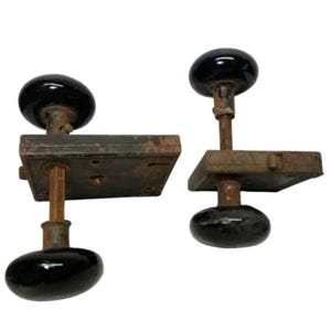 May include: Two vintage door handle sets featuring smooth, round, black knobs attached to rectangular, weathered, rusted metal backplates. Each assembly includes a central metal spindle connecting the knobs through the mounting plate hardware.