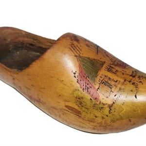 May include: Wooden clog with a painted landscape design. The clog is made of light brown wood and features a carved-out interior. The painted design includes green, red, and brown colors, depicting a building and foliage.