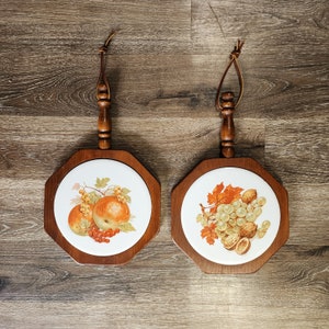May include: Two wooden trivets with a brown finish and a white ceramic center. One trivet features a design of apples and red berries, while the other features a design of grapes and walnuts.