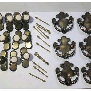 May include: A collection of hardware including twelve brass-toned metal knobs with white circular ceramic centers, six ornate brass-toned metal drop pulls with matching backplates, and several thin metal mounting bolts for installation.