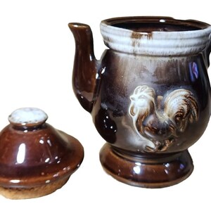 May include: A brown ceramic teapot with a rooster design and a matching lid. The teapot has a white glaze on the rim and a brown glaze on the body.