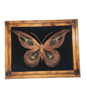 May include: A string art butterfly design created with copper and light yellow thread on a black background, encased in a rectangular wooden frame with a distressed, burnt-wood finish. The butterfly wings feature intricate geometric patterns.