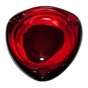 May include: A vintage ashtray with a deep red interior and a black exterior. The ashtray has a triangular shape with a rounded edge and a small notch for resting cigarettes. The glass is glossy and reflects light.