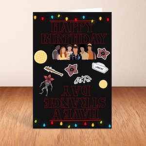 May include: A black birthday card with red text reading "HAPPY BIRTHDAY" and "HAVE A STRANGE DAY". The card features character illustrations, waffles, and themed graphics. Colourful string lights border the top and bottom.