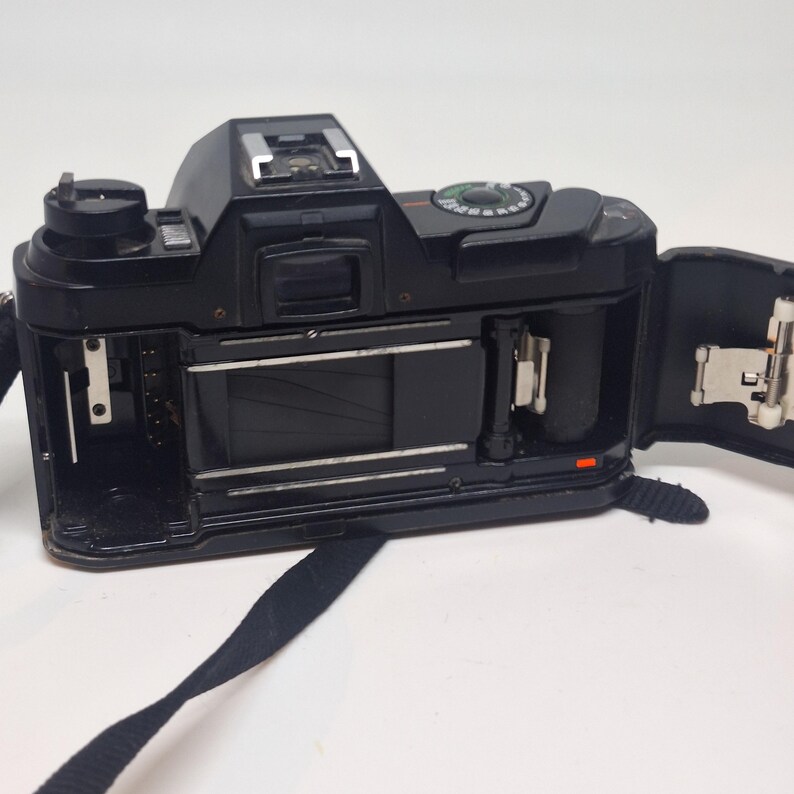 Puede incluir: C&aacute;mara de pel&iacute;cula vintage negra con la parte trasera abierta, revelando los componentes internos. La c&aacute;mara tiene una correa negra. El dise&ntilde;o incluye una montura para flash y diales. La c&aacute;mara est&aacute; sobre una superficie blanca. Mide unos 14 cm de largo.