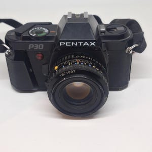Puede incluir: C&aacute;mara Pentax P30 negra con lente y correa. La c&aacute;mara tiene la palabra "PENTAX" impresa en la parte delantera. La lente tiene el n&uacute;mero "1871097" impreso. La correa de la c&aacute;mara es negra.