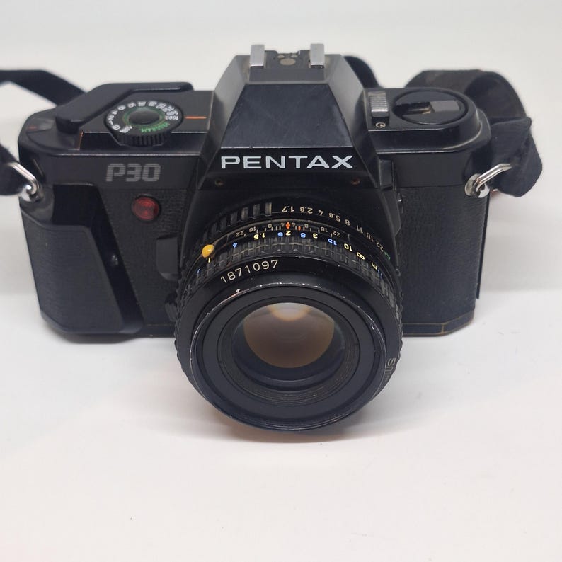 Puede incluir: C&aacute;mara Pentax P30 negra con lente y correa. La c&aacute;mara tiene la palabra "PENTAX" impresa en la parte delantera. La lente tiene el n&uacute;mero "1871097" impreso. La correa de la c&aacute;mara es negra.