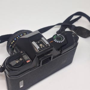 Puede incluir: C&aacute;mara de pel&iacute;cula vintage negra con lente y correa negra. La c&aacute;mara tiene un agarre texturizado y varios diales y botones. La lente tiene marcas para varias distancias focales. La c&aacute;mara est&aacute; sobre una superficie blanca.