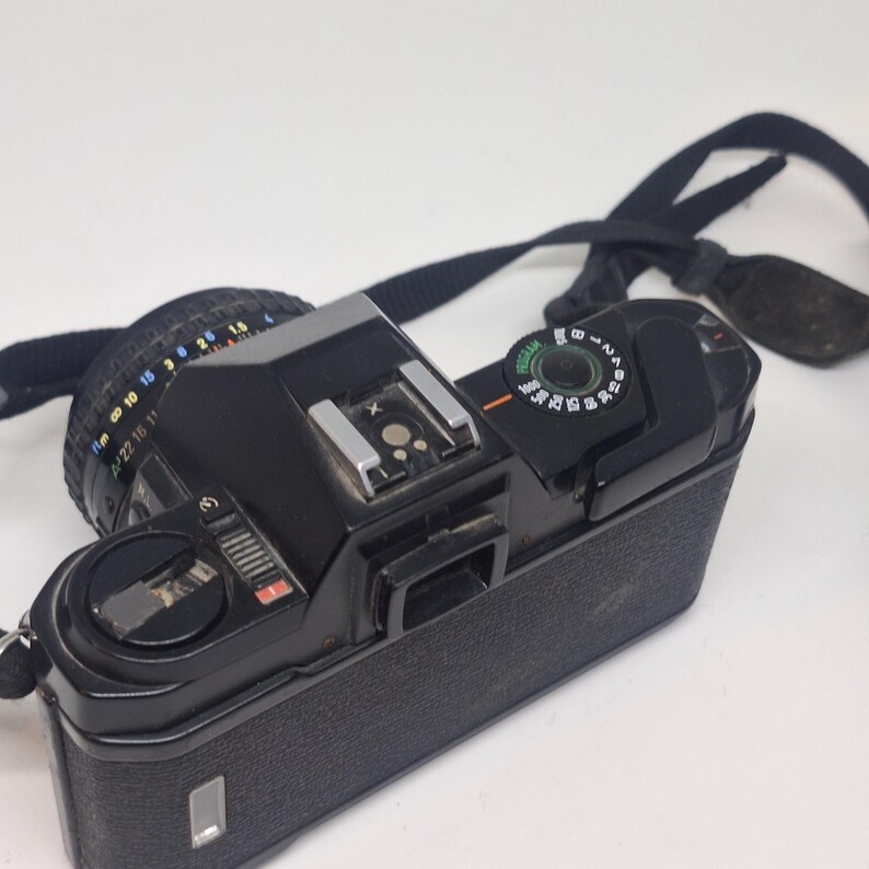 Puede incluir: C&aacute;mara de pel&iacute;cula vintage negra con lente y correa negra. La c&aacute;mara tiene un agarre texturizado y varios diales y botones. La lente tiene marcas para varias distancias focales. La c&aacute;mara est&aacute; sobre una superficie blanca.