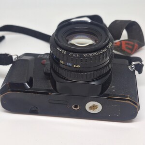 Puede incluir: C&aacute;mara Pentax P30 negra con una lente de 50 mm. La lente est&aacute; marcada SMC Pentax-A 1:1.7. La correa de la c&aacute;mara es negra con un dise&ntilde;o rojo y blanco. La c&aacute;mara est&aacute; sobre una superficie blanca.