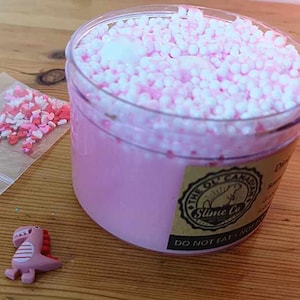 Op de afbeelding: Een doorzichtige plastic container gevuld met roze slijm en witte en roze kralen. Een kleine zak met hartvormige hagelslag en een roze dinosaurusbeeldje zijn ook aanwezig. De container heeft een label met "The Oil Canadian Slime Co."