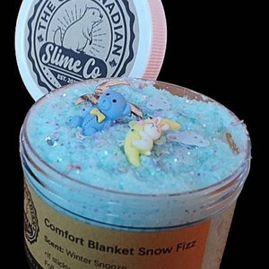 May include: A blue slime with glitter and small plastic charms in a clear jar with a brown label that reads "Comfort Blanket Snow Fizz" and "Scent: Winter Snooze". The label also includes instructions for use and a warning that the slime is not food.