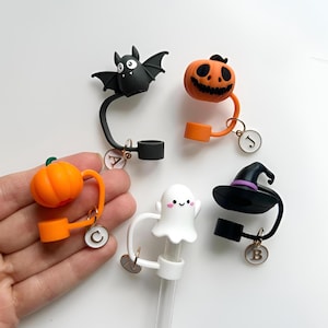 May include: A selection of Halloween-themed straw toppers. The toppers feature a black bat, an orange pumpkin, a white ghost, a witch's hat, and another orange pumpkin. Each topper has a charm with a letter.