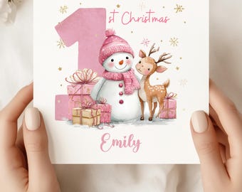 May include: A pink and white Christmas card featuring a snowman, a deer, and wrapped gifts. The card reads "1st Christmas" and "Emily" in pink script. Gold star accents complete the festive design.