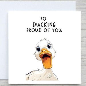 Peut inclure: Carte de vœux blanche avec une illustration de canard de dessin animé. Le canard a un bec jaune et regarde vers le haut avec une expression surprise. Le texte "SO DUCKING PROUD OF YOU" est écrit au-dessus du canard.