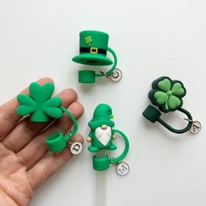 May include: Four green silicone straw toppers with charms. The toppers feature a leprechaun, a shamrock, a hat, and a shamrock. The charms are gold and have the letters C, D, A, and M.