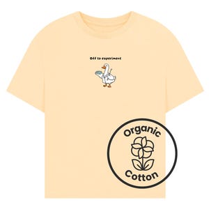 May include: A light yellow t-shirt featuring a small graphic of a white duck wearing a lab coat and carrying a blue petri dish, with the text "Off to experiment" above it. A circular black logo with a flower icon and the text "Organic Cotton" is visible.