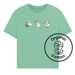 May include: A light green t-shirt featuring a graphic of three white geese dressed as scientists performing laboratory tasks. A circular black and white logo on the lower right displays a flower icon with the text Organic Cotton.
