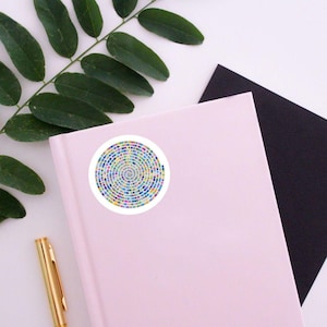 May include: A pink notebook with a white sticker featuring a colorful spiral design. A gold pen, a black notepad, and green leaves are also visible on a white surface.