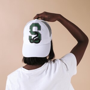May include: White trucker hat with a green and grey letter 'S' with a black snake inside. The snake is coiled and has its head raised.
