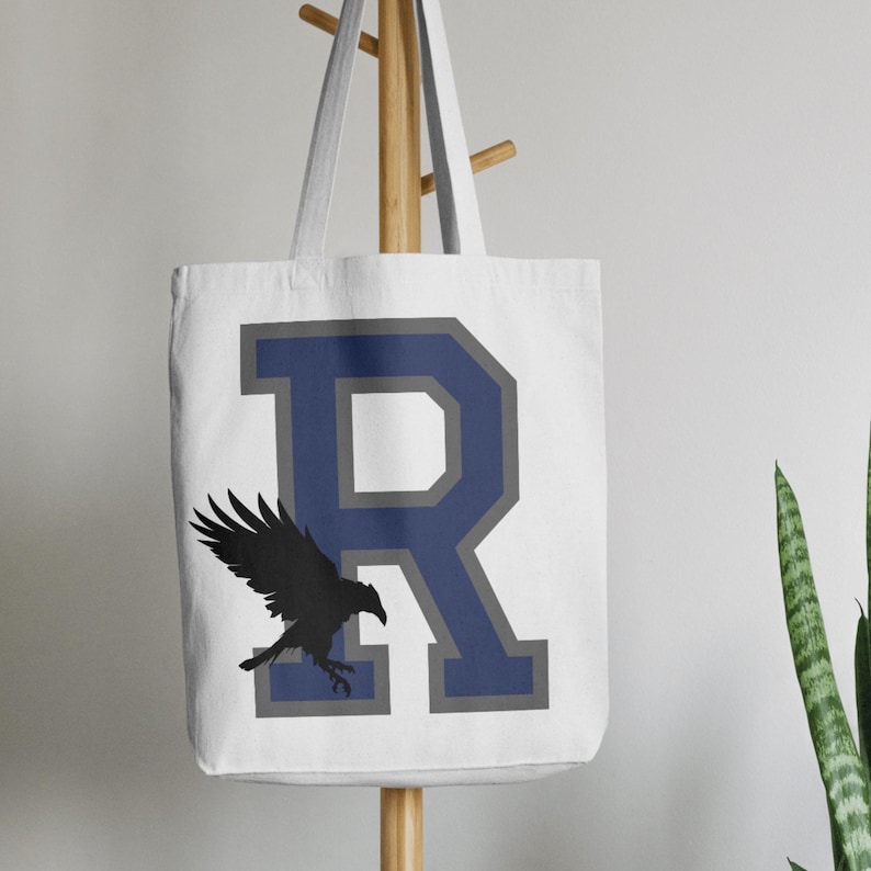 May include: White canvas tote bag featuring a navy blue and grey letter "R" with a black silhouette of a raven flying in front of it.