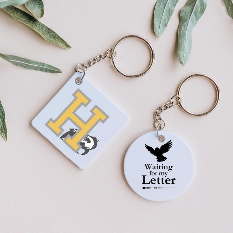 May include: Two keychains with white circular and square charms. The square charm has a yellow "H" with a black badger on it. The circular charm has a black silhouette of a bird with the text "Waiting for my Letter" and a wand.
