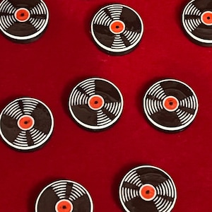 May include: A collection of ten circular buttons designed to resemble miniature vinyl records. Each button features a black surface with white concentric grooves and a small, bright orange circular center, arranged in a scattered pattern.