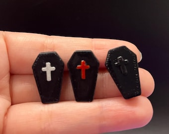 Puede incluir: Tres pequeños pines con forma de ataúd negro. El primero presenta una cruz blanca, el segundo una cruz roja y el tercero una cruz negra. Cada ataúd tiene un borde texturizado y un acabado suave y brillante en los detalles de las cruces.