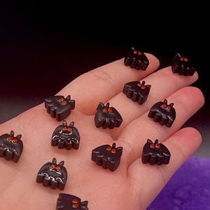 May include: A collection of small, black, bat-shaped beads or charms. Each piece features two pointed ears and two prominent, bright orange eyes. Some of the bat figures have additional white painted details on their bodies, while others are plain black.