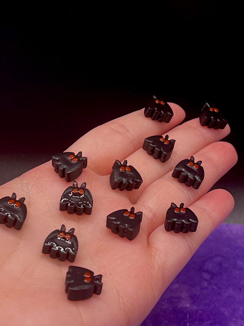 May include: A collection of small, black, bat-shaped beads or charms. Each piece features two pointed ears and two prominent, bright orange eyes. Some of the bat figures have additional white painted details on their bodies, while others are plain black.