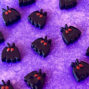 May include: A collection of small, black moth-shaped charms with two prominent red eyes. Each charm features a smooth, glossy finish and a distinct silhouette with antennae at the top and a scalloped bottom edge, arranged in a scattered pattern.