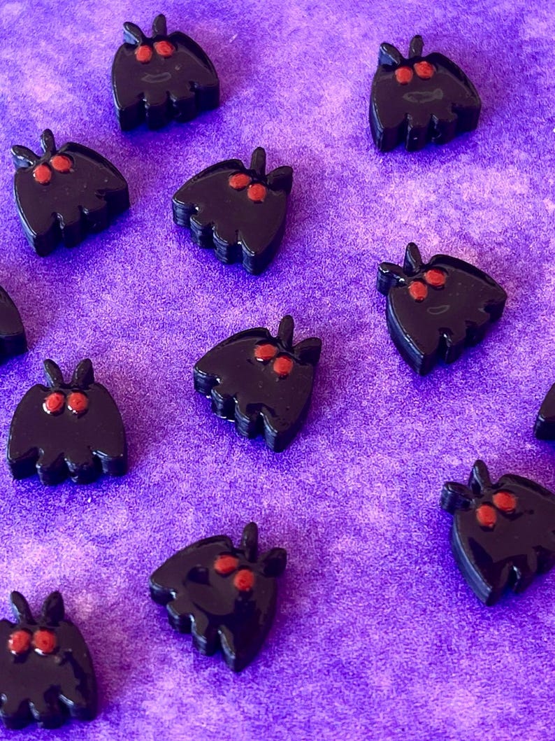 May include: A collection of small, black moth-shaped charms with two prominent red eyes. Each charm features a smooth, glossy finish and a distinct silhouette with antennae at the top and a scalloped bottom edge, arranged in a scattered pattern.