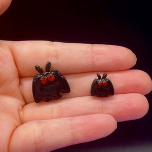 May include: Two small, glossy black moth-shaped charms with prominent, rounded orange eyes and short antennae. The charms feature a smooth, reflective surface and are shaped like stylized insects with wings spread, one being larger than the other.