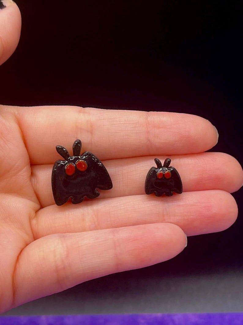 May include: Two small, glossy black moth-shaped charms with prominent, rounded orange eyes and short antennae. The charms feature a smooth, reflective surface and are shaped like stylized insects with wings spread, one being larger than the other.