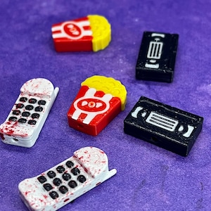 May include: Five colorful 3D printed charms, including a red and yellow popcorn bucket, a black VHS tape, and two white phones with red splatters.