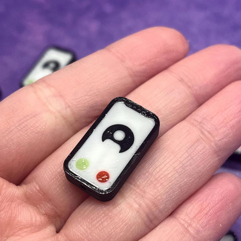 Puede incluir: Un peque&ntilde;o dije rectangular con borde negro y centro blanco. El centro presenta un icono de luna creciente negra sobre un peque&ntilde;o c&iacute;rculo verde claro y un peque&ntilde;o c&iacute;rculo rojo. El dije tiene un acabado suave, brillante y esquinas redondeadas.