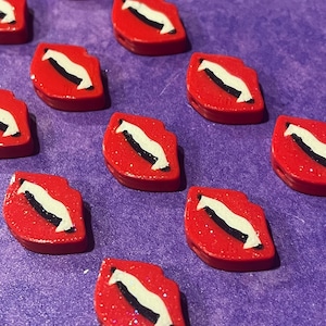 May include: Red lip-shaped embellishments with a white and black design. The embellishments are scattered across a purple surface. The red has a glittery finish, adding a touch of sparkle to the design.