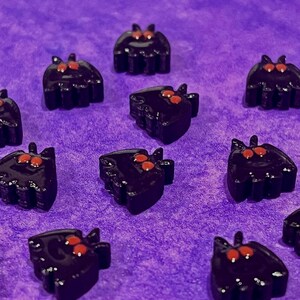 May include: A collection of small, black, bat-shaped charms with two prominent red eyes. Each charm features a smooth, glossy finish and a distinct silhouette with pointed ears and rounded wings, arranged in a scattered pattern across the frame.