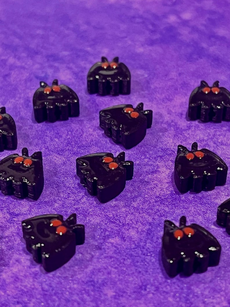 May include: A collection of small, black, bat-shaped charms with two prominent red eyes. Each charm features a smooth, glossy finish and a distinct silhouette with pointed ears and rounded wings, arranged in a scattered pattern across the frame.