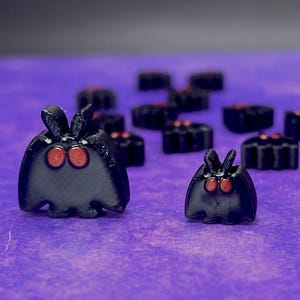 May include: A collection of small, black, moth-shaped figurines with two prominent red eyes and upright antennae. The figures vary in size, with two larger ones in the foreground and several smaller ones arranged in the background.