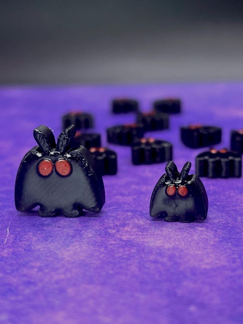 May include: A collection of small, black, moth-shaped figurines with two prominent red eyes and upright antennae. The figures vary in size, with two larger ones in the foreground and several smaller ones arranged in the background.