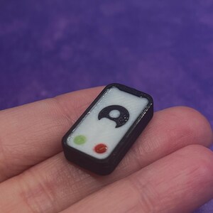 Puede incluir: Un peque&ntilde;o dije rectangular con borde negro y centro blanco. La cara muestra un icono de luna creciente negra posicionado sobre un peque&ntilde;o c&iacute;rculo verde y un peque&ntilde;o c&iacute;rculo rojo, asemej&aacute;ndose a una interfaz simplificada o un bot&oacute;n.