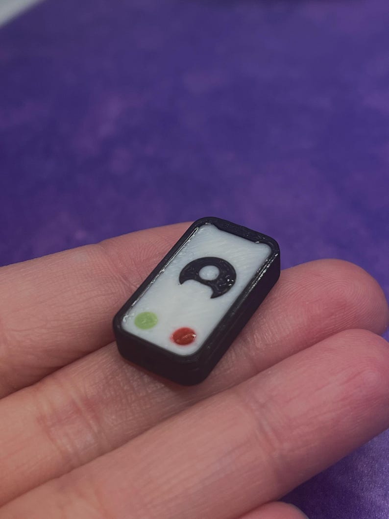 Puede incluir: Un peque&ntilde;o dije rectangular con borde negro y centro blanco. La cara muestra un icono de luna creciente negra posicionado sobre un peque&ntilde;o c&iacute;rculo verde y un peque&ntilde;o c&iacute;rculo rojo, asemej&aacute;ndose a una interfaz simplificada o un bot&oacute;n.