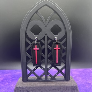Può includere: Un paio di orecchini pendenti con ciondoli neri a forma di bara con una croce rossa centrale. Gli orecchini sono appesi a perni in metallo color argento e sono esposti su un supporto nero a forma di arco con motivo a griglia.