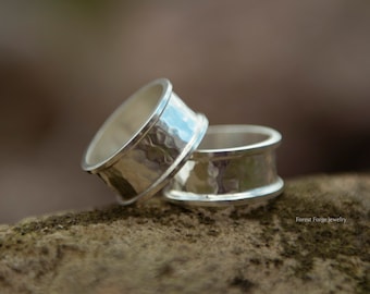May include: Two silver rings with a hammered texture are stacked on a stone. The rings have a wide band and a polished edge. The background is blurred, with a natural, earthy color palette.