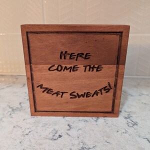 May include: A wooden block with the phrase "HERE COME THE MEAT SWEATS!" engraved in black. The block is a warm brown color and has a framed border. The text is centered and in a handwritten style.