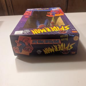 May include: A red and blue Spider-Man action figure in a box. The box is red, blue, and yellow with the words "Deluxe Edition Super Posable Spider-Man" on the front. The figure is from the "The New Animated Series" and is a "Toy Biz" product.