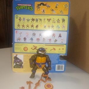 May include: Teenage Mutant Ninja Turtles action figure of Donatello, the purple masked turtle, with his signature bo staff, sai, and katanas. The figure is posed in a fighting stance with a brown and orange shell and a purple bandana. The figure is packaged on a blue and yellow card with the Teenage Mutant Ninja Turtles logo and the text "Collect all the Wacky Action Turtles!" and "We're looking for a few good mutants!"