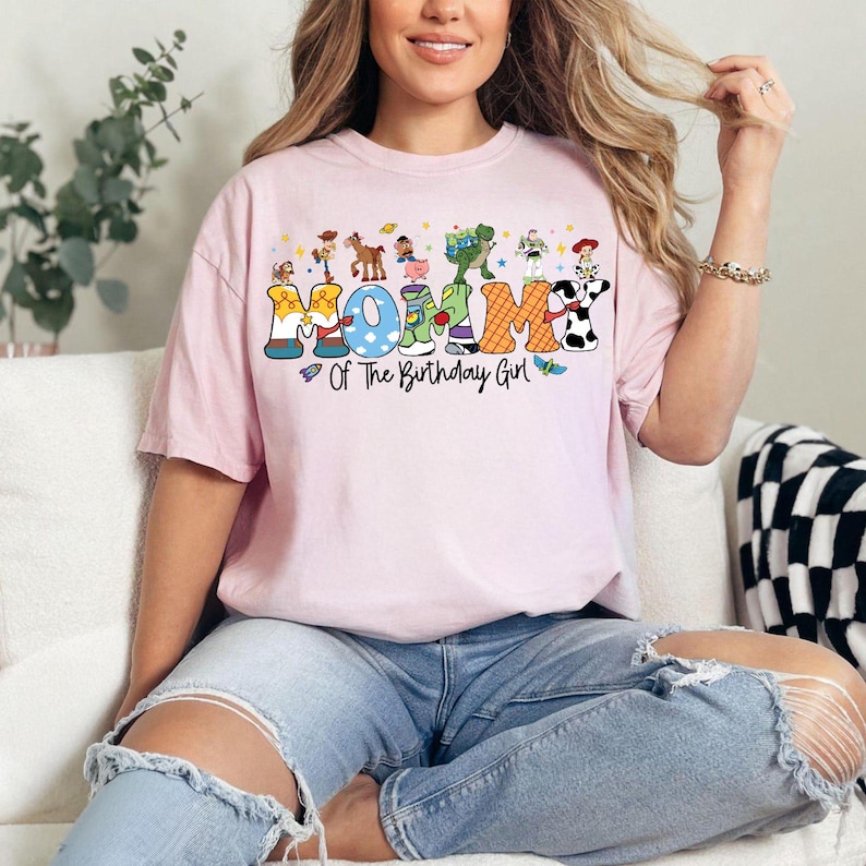 May include: A light pink t-shirt with the word "MOM" in colorful block letters, each letter decorated with cartoon characters. The text "Of The Birthday Girl" is below the word "MOM".