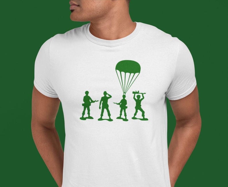 Peut inclure: T-shirt blanc avec un motif vert de quatre soldats en plastique et un parachute. Les soldats tiennent des fusils jouets et le parachute est ouvert.